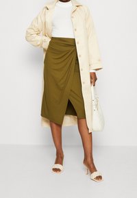 Beige Trenchcoat, weißes enges Oberteil, olivgrüne, drapierte Rock, hält eine weiße Handtasche und trägt beige High Heels mit geflochtenen Details.