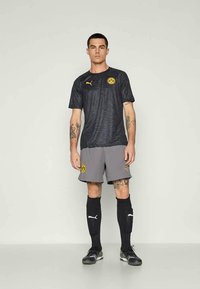 Muž v čiernom vzorovanom futbalovom drese Borussia Dortmund, s šedými šortkami, čiernymi ponožkami Puma a čiernymi kopačkami, stojí na jednofarbnom pozadí.