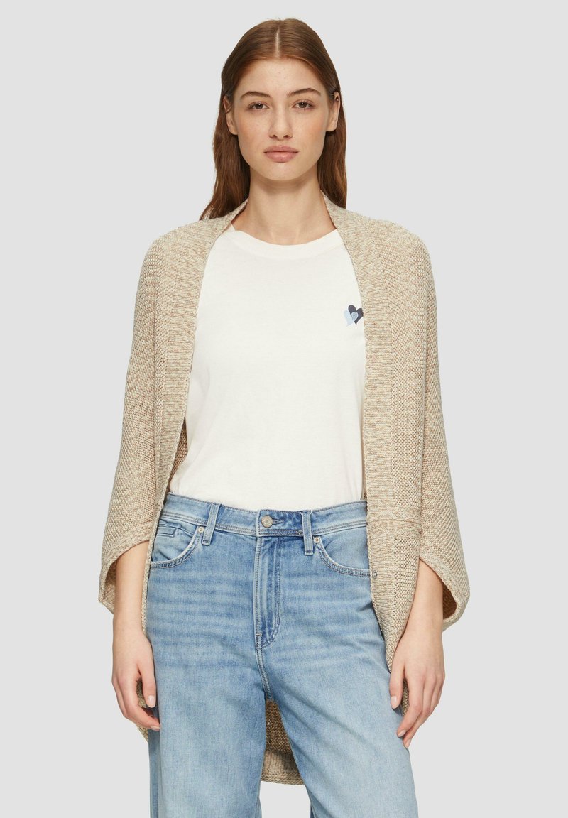 Beige gestrickte Cardigan mit offener Vorderseite, strukturierter Stoff, getragen über einem weißen T-Shirt mit einem blauen Herzlogo. Hellblaue Jeans vervollständigen das Outfit.