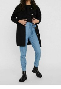 Abrigo largo negro con dos bolsillos en el pecho, combinado con pantalones azul claro con una cintura atada. Llevado con botas negras de suela gruesa.