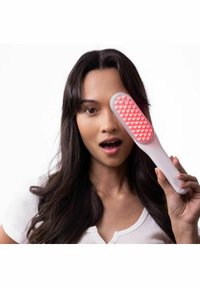 Jeune femme aux longs cheveux noirs tient une brosse de massage rose devant un œil, la bouche légèrement ouverte, sur fond blanc.