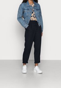 Kvinna som bär en blå denimjacka, topp med djurmotiv, svarta snörbyxor och vita sneakers, stående mot en enkl vit bakgrund.