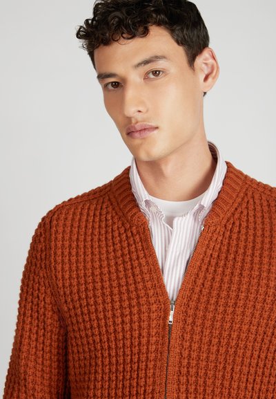Teksturowany kardigan w kolorze rdzy z zamkiem błyskawicznym i prążkowanym wzorem. Nosi się go na jasnej, w paski koszuli, co podkreśla warstwowy styl.
