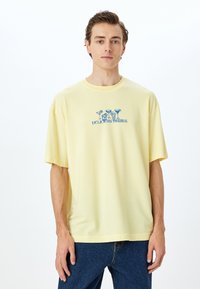 Camiseta de manga corta amarilla clara, hecha de algodón suave, que presenta un diseño gráfico azul de bebidas con el texto "BEBIDAS DELICIOSAS" debajo.