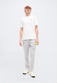 Νέος άνδρας που φοράει λευκό T-shirt, ανοιχτό γκρι φόρμες, κρεμ αθλητικά παπούτσια και απαλό ροζ καπέλο, κρατώντας ένα πράσινο μήλο στο δεξί του χέρι.