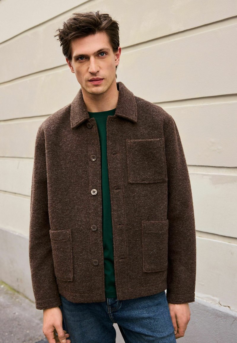 Veste en laine marron avec un col, fermeture à boutons, deux poches avant et une finition texturée, portée sur une chemise verte et un jean.