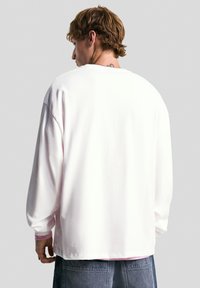 Un jeune homme aux cheveux châtain clair portant une chemise blanche à manches longues ample et un jean en denim sombre, se tenant contre un fond gris uni.