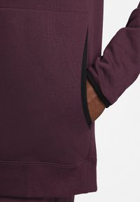 Haut bordeaux à manches longues avec une finition texturée, présentant une poche latérale noire et un poignet noir. Design élégant avec une coupe décontractée.