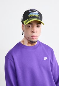 Fekete sapka élénk sárga peremmel, merész grafikákkal; lila pulóver fehér Nike logóval, sima textúrával, laza szabással.