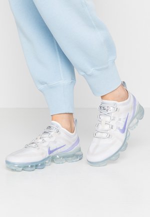Osoba nosząca białe i lawendowe buty Nike Air VaporMax w połączeniu z jasnoniebieskimi, podwiniętymi dresowymi spodniami na białej podłodze.