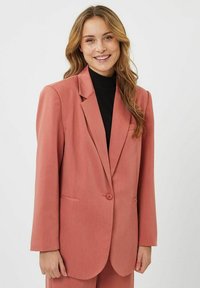 Blazer rosa claro en una tela texturizada, con solapa de muesca, cierre de un botón y dos bolsillos tipo deslizamiento. Estilizado sobre una blusa negra.