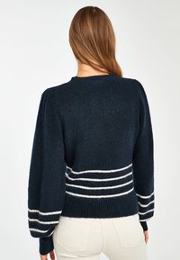 Pull tricoté bleu marine avec tissu texturé, manches ballon et rayures horizontales blanches à la taille. Encolure ronde, coupe courte.