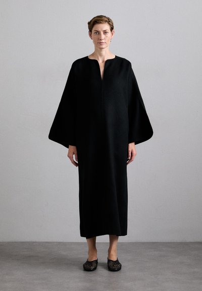 Femme debout avec une expression neutre portant une longue robe noire à manches larges et des chaussures tissées noires sur un sol gris.