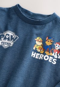 Tumši zils T-krekls ar "PAW Patrol" logotipu un krāsainiem trīs suņu karikatūru tēliem, uz tā ir vārds "HEROES" izcelts baltā treknā fontā.