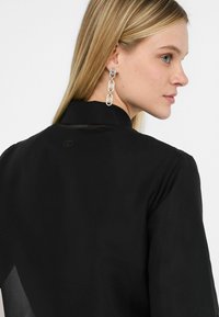 Blusa nera con colletto alto e dettagli trasparenti sulle maniche. Orecchini a catena argentati con accenti collegati. Branding minimalista sul collo.