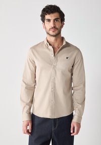 Camicia a maniche lunghe color beige chiaro con colletto a punta, con un piccolo logo blu navy sul petto e un tessuto liscio e texture.
