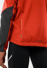 Giacca rossa con maniche nere, caratterizzata da una finitura testurizzata, accenti riflettenti e un logo sull'orlo. Pantaloni neri slim-fit con dettagli grigi.
