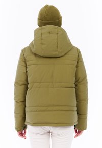 Persoon in een olijfgroene gewatteerde jas en bijpassende beanie, van achteren weergegeven, staand tegen een witte achtergrond.