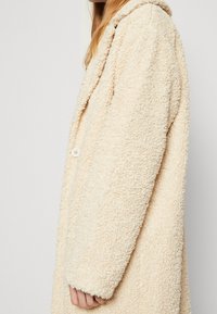 Cappotto in felpa beige con materiale testurizzato e soffice, colletto a rever e chiusura con un bottone. La vista laterale mette in evidenza la vestibilità rilassata e la superficie morbida.