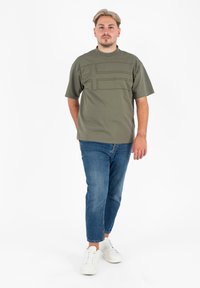 Oliwkowa koszula z krótkim rękawem w teksturowe poziome paski, w połączeniu z jasnoniebieskimi dżinsami i białymi trampkami. Gładkie tło.