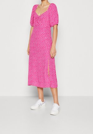 Pink midi dress with puff sleeves, white heart pattern, and a front slit. Model wearing white sneakers. Smooth fabric texture.