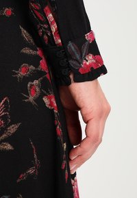 Tela de vestido negro con flores rojas y moradas. Manga con puño y cuatro botones negros, con un borde con volantes, que muestra una textura suave.