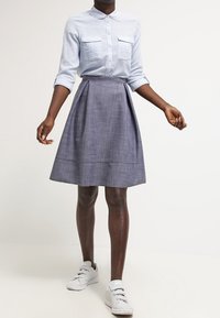 Camisa de botones azul claro con dos bolsillos en el pecho, combinada con una falda gris con pliegues hasta la rodilla y zapatillas blancas.