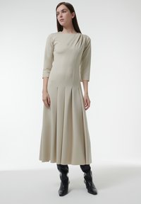 Vestido beige de manga larga con escote redondo, falda plisada y detalles laterales asimétricos; combinado con botas negras hasta la rodilla.