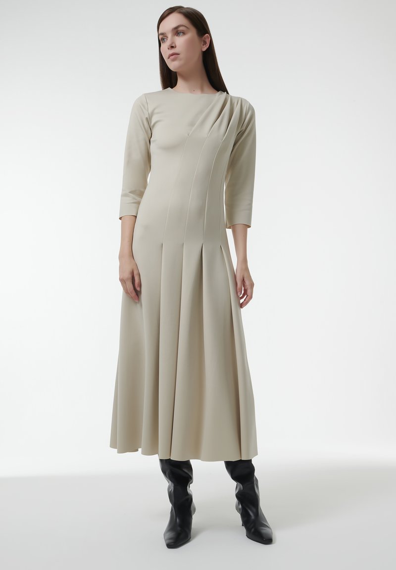 Vestido beige de manga larga con escote redondo, falda plisada y detalles laterales asimétricos; combinado con botas negras hasta la rodilla.