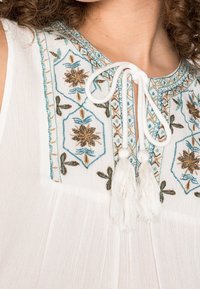 Blouse blanche ornée de délicates broderies bleues et brunes, avec un nœud au cou agrémenté de franges, et un design ample et léger.