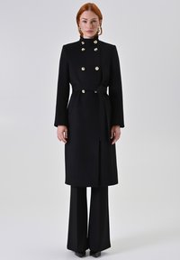 Zwarte wollen jas met gouden knopen, hoge kraag, ceintuur in de taille en lange mouwen. Draagt over zwarte wijde broekspijpen. Glad textuur.
