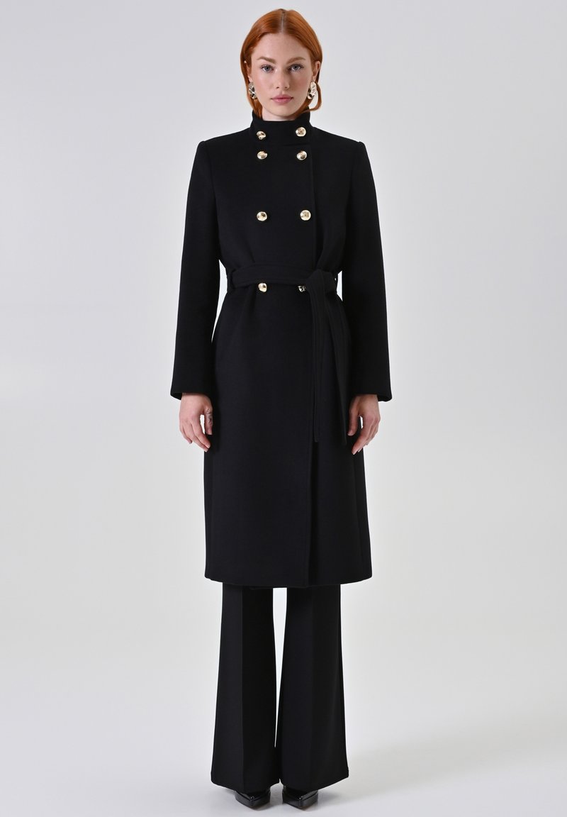 Zwarte wollen jas met gouden knopen, hoge kraag, ceintuur in de taille en lange mouwen. Draagt over zwarte wijde broekspijpen. Glad textuur.