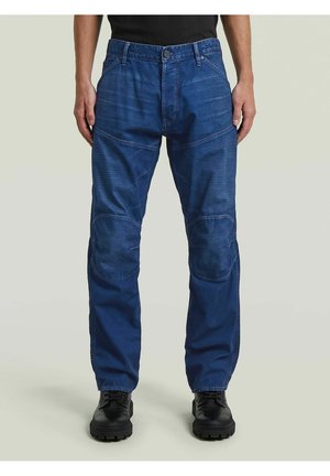 Mann trägt blaue Jeanshosen mit Knieschonern und schwarzen Schuhen, steht vor einem schlichten hellgrauen Hintergrund.