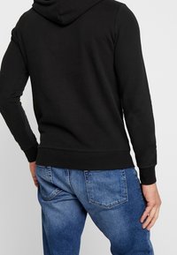 Mężczyzna w czarnej bluzie z kapturem i niebieskich dżinsach stoi plecami do kamery na jednolitym tle.