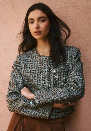 Veste texturée courte en gris et marron atténués avec des sequins, dotée de deux poches poitrine et de boutons arrondis, soulignée d'un liseré contrastant.