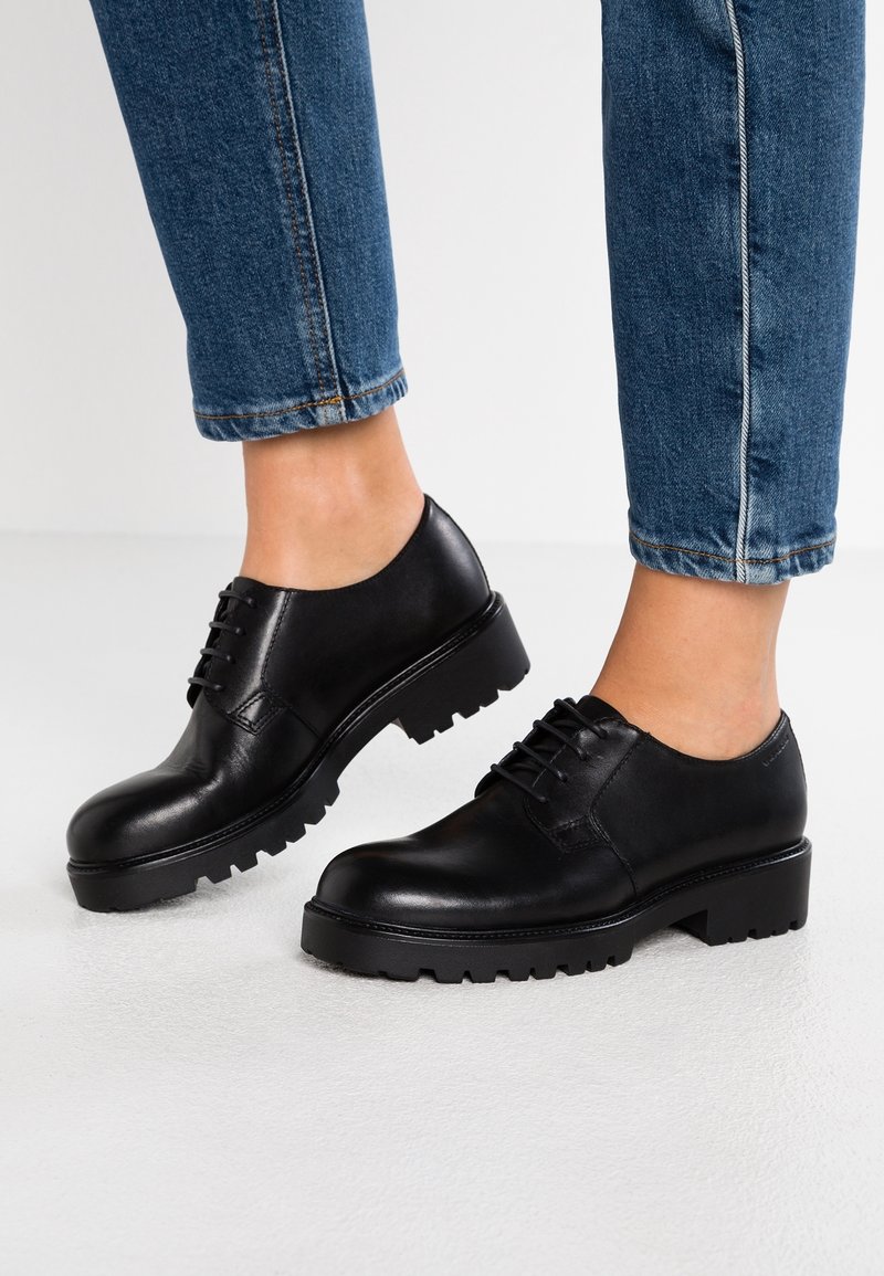 Chaussures en cuir noir avec embout rond, design à lacets et semelle en caoutchouc épaisse avec une bande de roulement texturée. Portées avec un jean bleu retroussé.