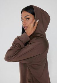 Bruine hoodie van zacht materiaal, met een aansluitend ontwerp, geribbelde mouwen en een capuchon met trekkoord. Glad textuur en minimale detailing.