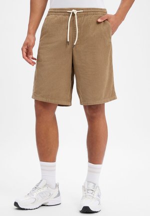 Uomo che indossa pantaloncini di velluto a coste beige con coulisse bianca, calzini bianchi e scarpe da ginnastica bianche, in piedi con una mano in tasca.