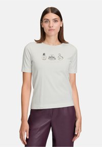 Femme portant un haut gris clair à manches courtes avec des illustrations de flacons de parfum et du texte, associé à un pantalon en cuir violet, sur un fond uni.