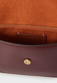 Burgunderfarbene Lederhandtasche mit weichem Innenfutter, Reißverschlussverschluss, goldfarbenen Beschlägen und einem sichtbaren Markenschild, das auf einem braunen Hintergrund geprägt ist.