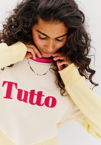 Mikina s růžovým logem "Tutto", smetanovým a žlutým designem. Má žebrovaný výstřih a dlouhé rukávy s kontrastními manžetami.