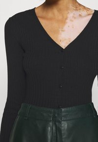 Schwarze gerippte Langarm-Cardigan mit V-Ausschnitt und sechs vorderen Knöpfen, kombiniert mit dunkelgrünen, hoch taillierten Lederhosen.