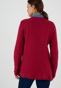 Cárdigan de manga larga en tejido de punto rojo oscuro, con un patrón texturizado y un corte relajado, que se lleva sobre una camisa azul por debajo.