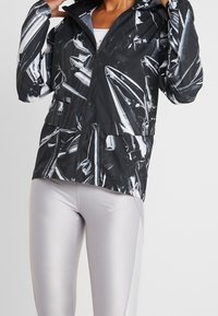 Black jacket with a shiny, abstract silver pattern, featuring a zip front, hood, and two front pockets; paired with grey leggings.