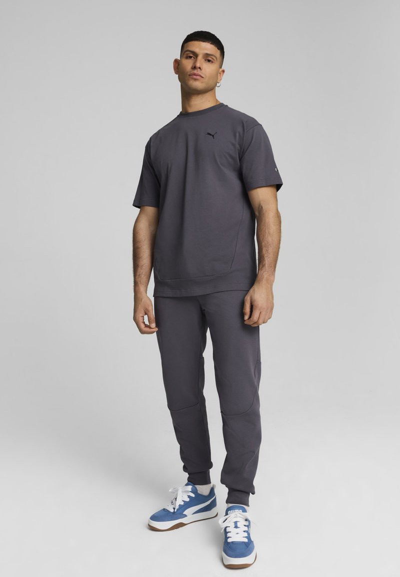 Grijze sport T-shirt en bijpassende joggers met zakken; stof lijkt zacht; blauwe en witte sneakers met bruine accenten. Zwart logo op de borst.