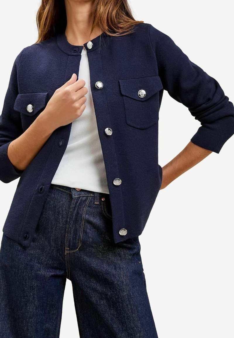 Femme portant un cardigan bleu marine avec des boutons argentés sur un haut blanc, associé à un jean bleu foncé, posant avec une main près de la poitrine et l'autre sur la hanche.
