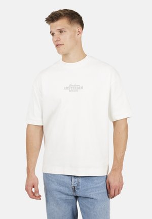 Giovane uomo che indossa una camicia bianca a maniche corte con la scritta "Amsterdam Est. 1972", abbinata a jeans azzurri, che guarda leggermente verso sinistra.