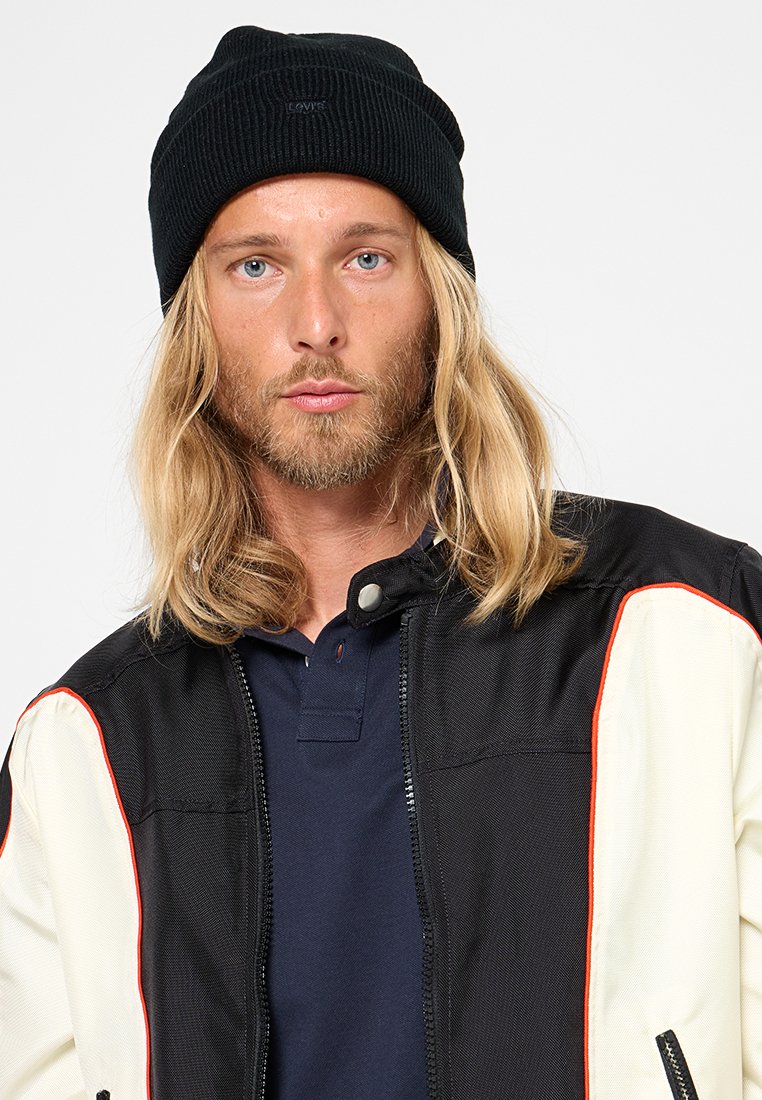 Bonnet côtelé noir, coupe décontractée. Le modèle porte une veste noire et crème avec des accents rouges par-dessus un polo bleu marine. Cheveux longs blonds visibles.