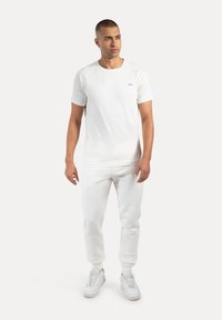 Mann steht mit entspannten Händen, trägt ein komplett weißes Outfit mit einem kurzärmeligen Hemd, Jogginghosen und weißen Turnschuhen vor einem weißen Hintergrund.