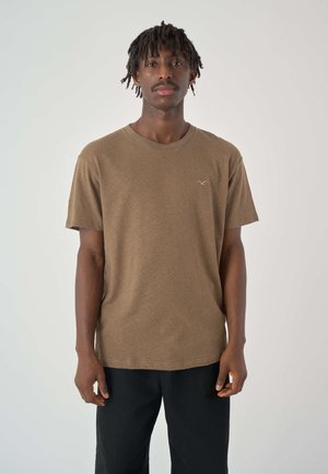 Braunes Baumwoll-T-Shirt mit rundem Hals und kurzen Ärmeln, das ein dezentes Logo auf der Brust und eine lockere Passform aufweist. Schwarze Hose wird getragen.